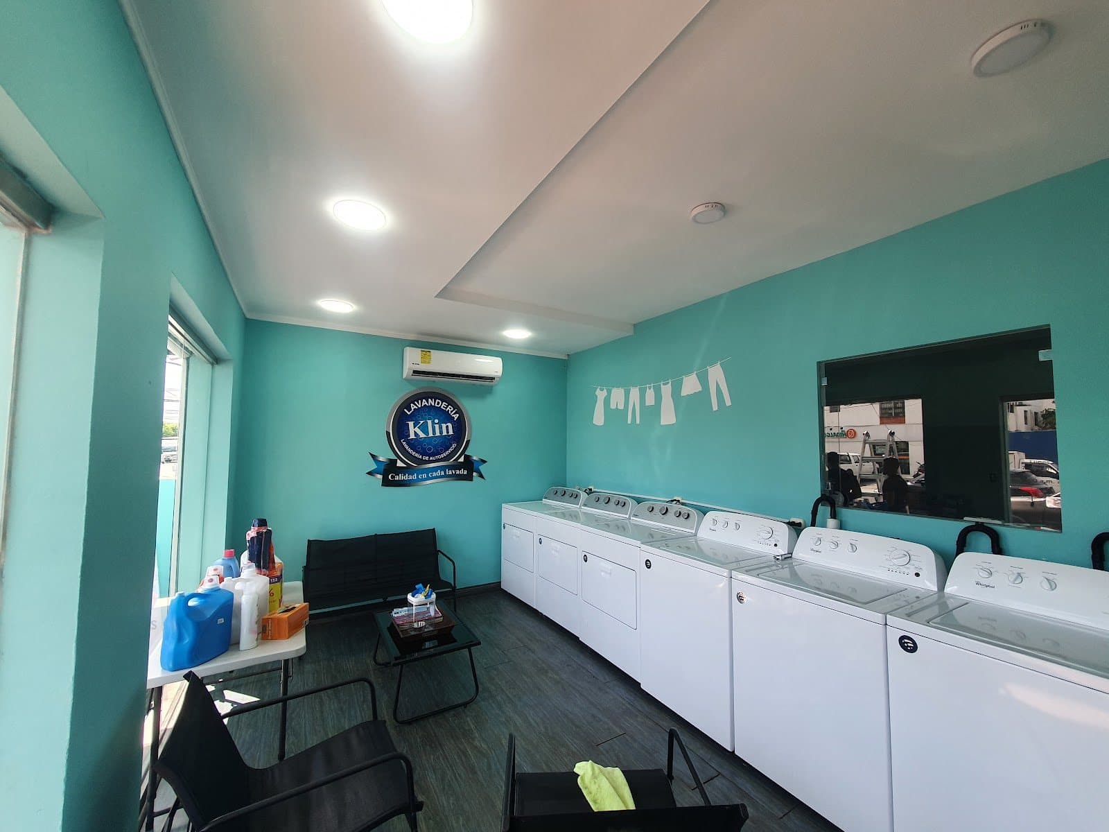 Interior de Lavanderia Klin en Los Prados con lavadoras y secadoras blancas, sillas de espera y el logo en la pared